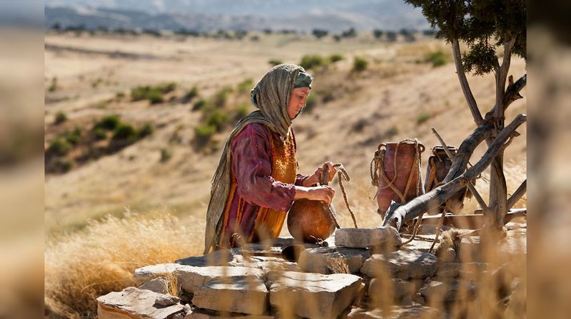 Kvinne med vannkrukker og lærsekk, som henter vann i en brønn omgitt av et tørt, solfylt landskap
