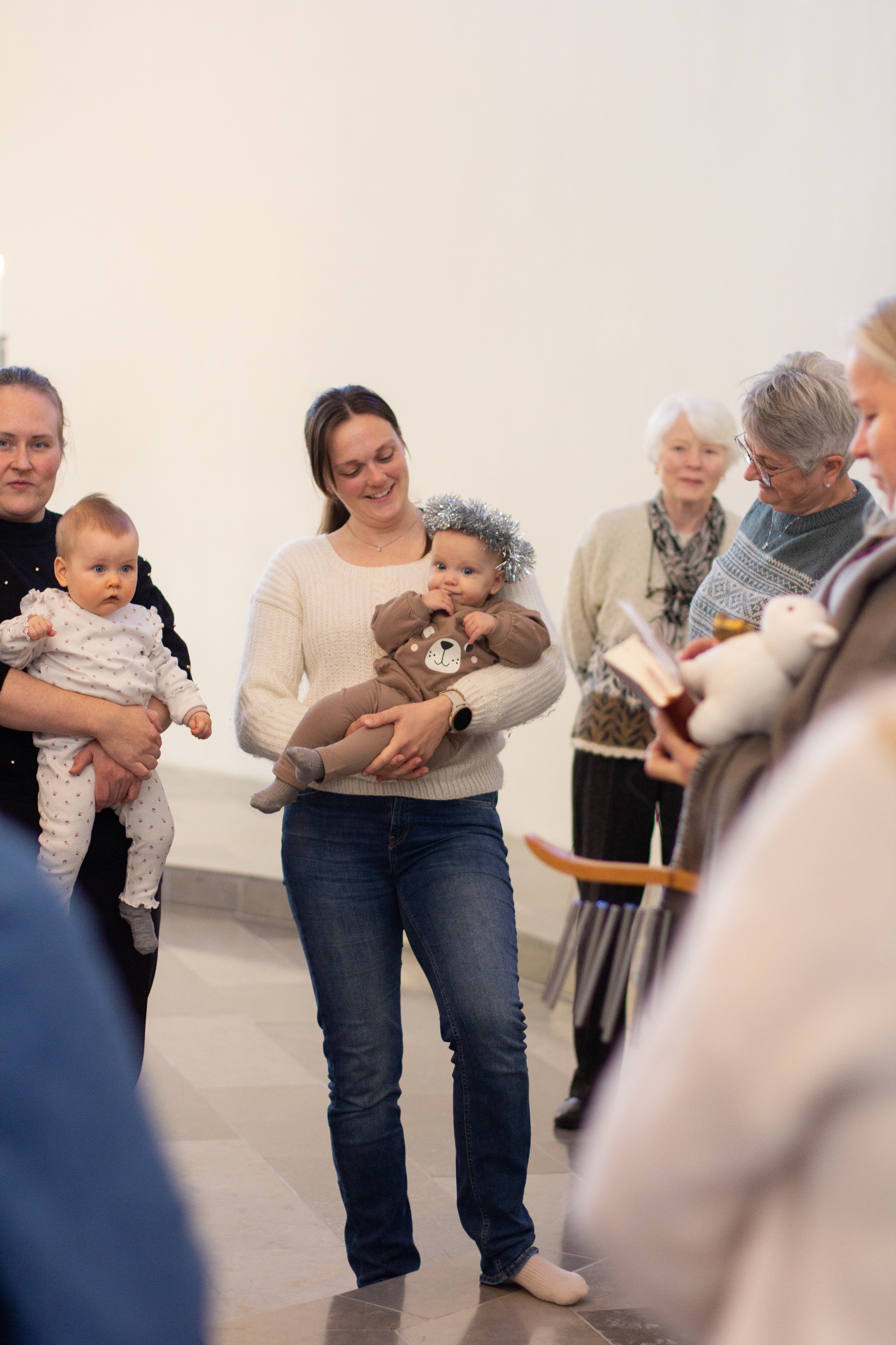 To mødre holder babyene sine, en baby har glitterkrans på hodet. På siden står noen bestemødre.